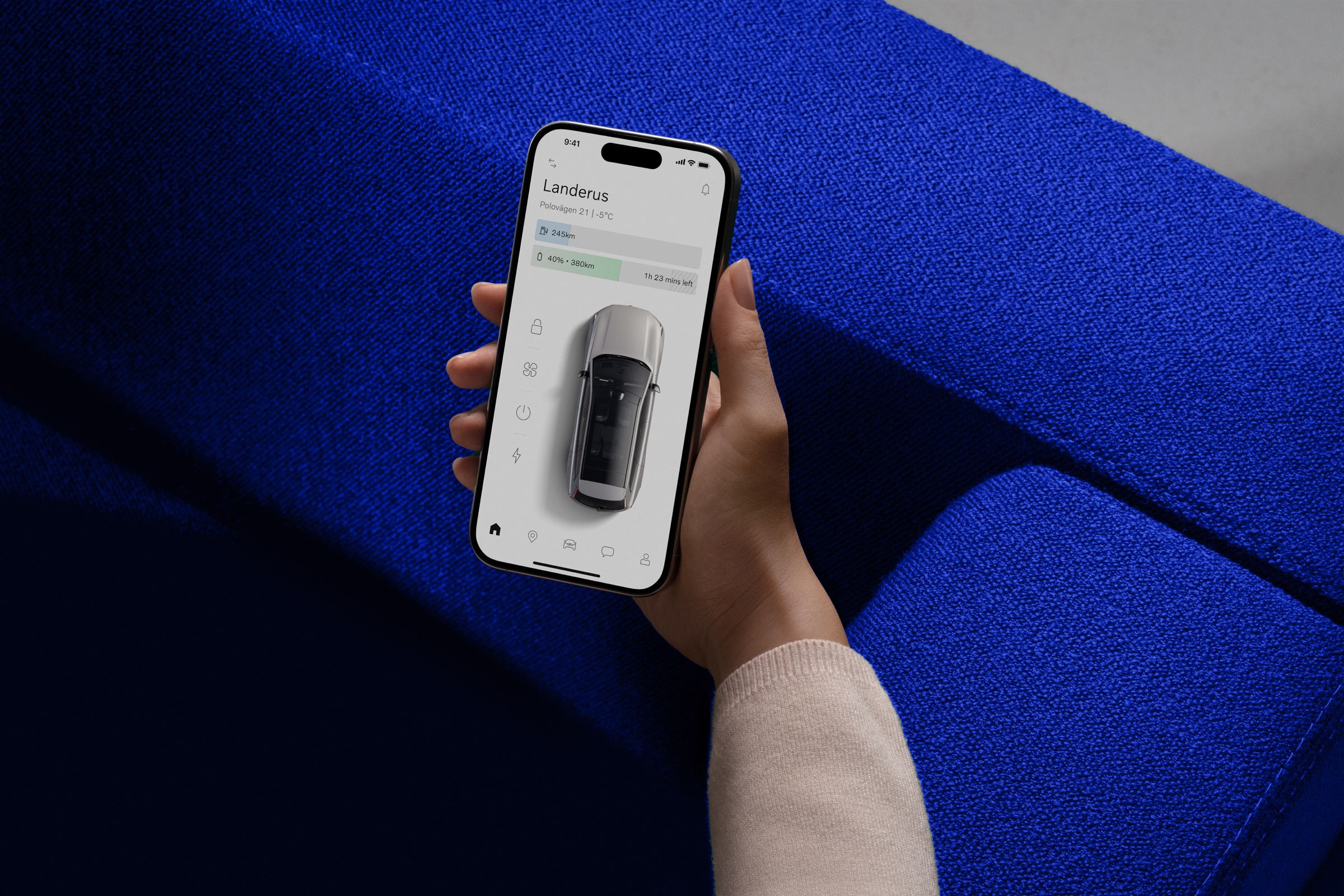 A hand holding a phone with the Volvo Cars app against a vivid blue sofa.